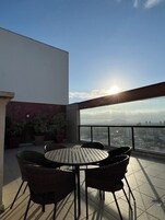 Una terraza en la azotea con una mesa redonda, sillas de mimbre, plantas en macetas y vistas a la ciudad.