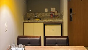 Uma cozinha moderna com bancada de granito, forno embutido e pia. Há duas luminárias pendentes amarelas, uma porta de madeira e uma mesa de jantar com quatro cadeiras.