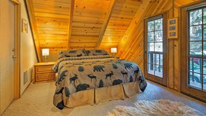 Een slaapkamer met een houten plafond, een bed met een deken met beertjes erop, twee nachtkastjes met lampen, en grote ramen met uitzicht op de omgeving.
