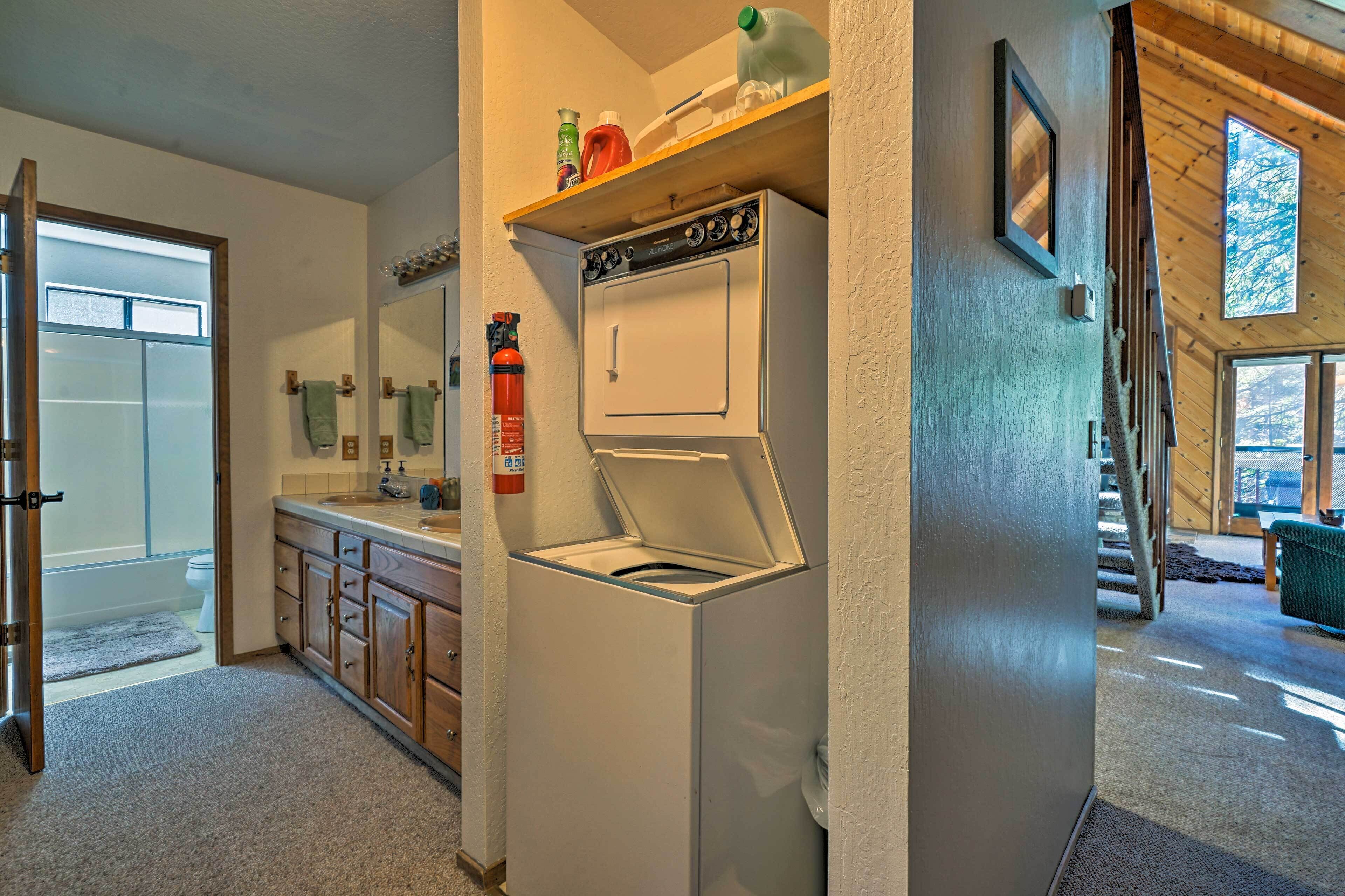 A laundry area with a front-loading washer and dryer, wooden cabinets, a small shelf with cleaning supplies, and a visible fire extinguisher.