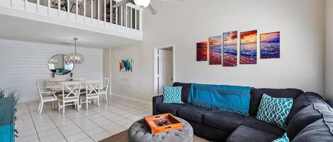 A spacious living room with a dark sofa, ottoman, and a dining area with a white table and chairs.