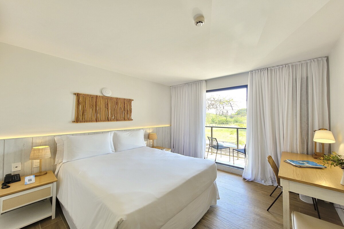 A hotel room with a large bed, bedside tables with lamps, a desk with a chair, and a view through the window.