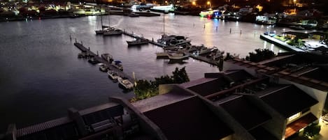 Un puerto deportivo con barcos amarrados, un edificio frente al agua con techo inclinado y una vista distante de un pueblo iluminado.