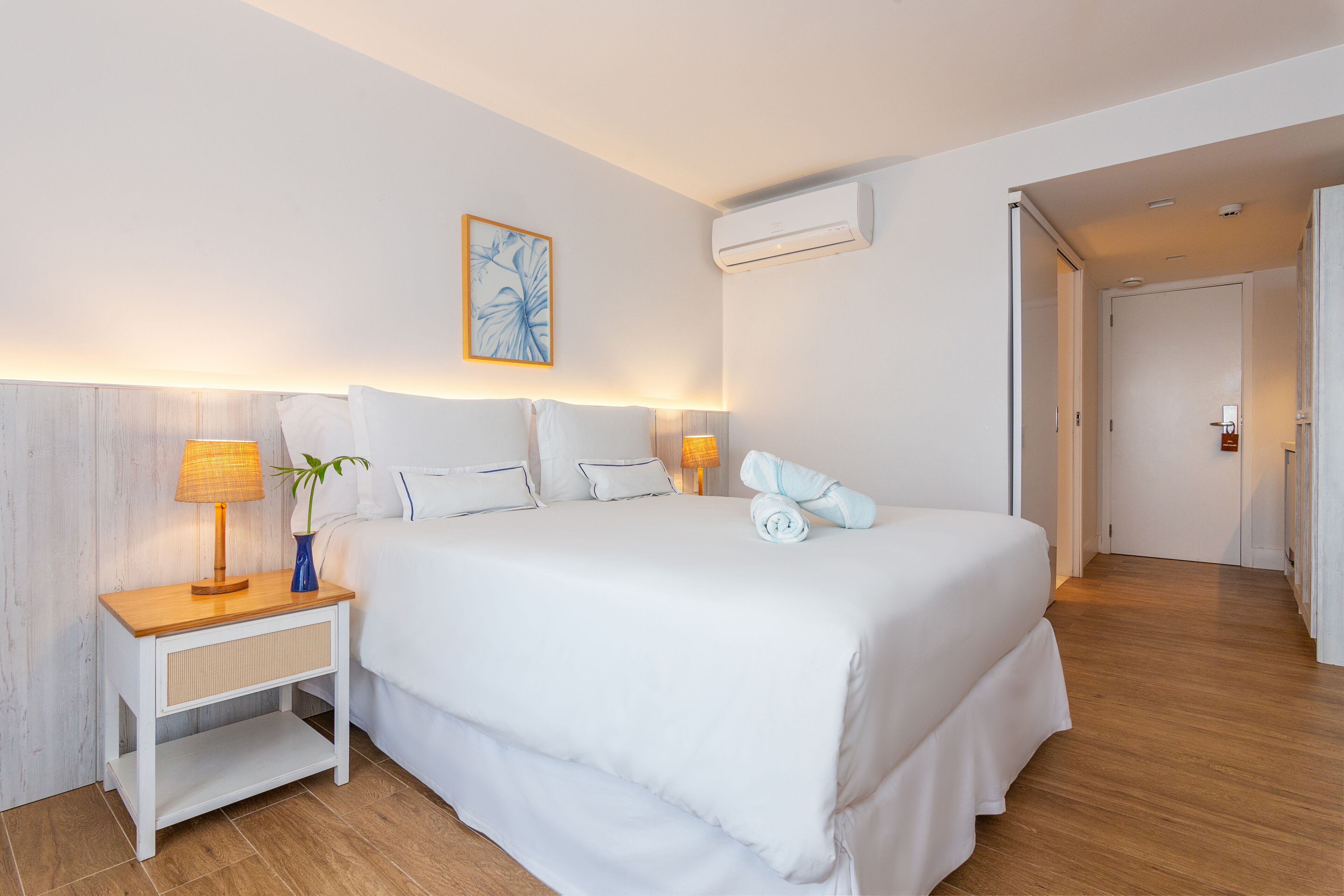 Habitación de hotel con una cama grande, lámparas de noche, aire acondicionado de pared y una puerta que da a otra habitación.