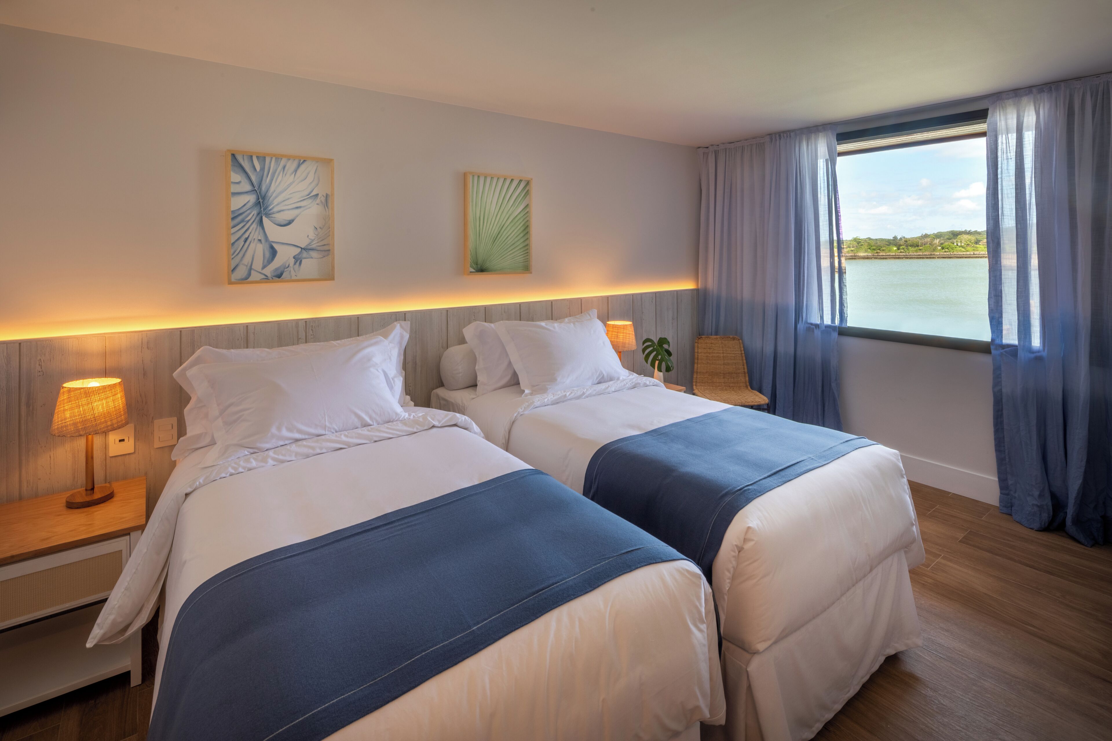 Et hotelværelse med to senge, et natbord med lampe, vægkunst og udsigt til vand.