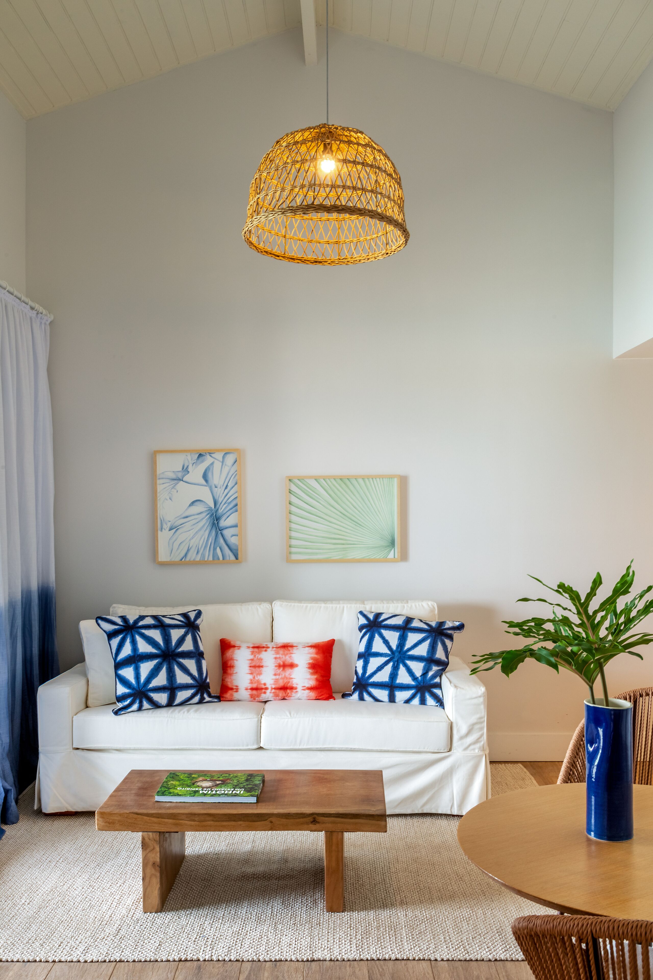 Un salon avec un canapé blanc, des coussins aux motifs bleus et rouges, une table basse en bois et un luminaire suspendu en rotin.