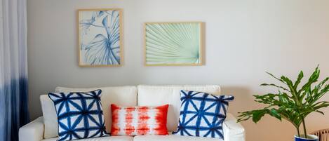 Un salon avec un canapé blanc, des coussins aux motifs bleus et rouges, une table basse en bois et un luminaire suspendu en rotin.