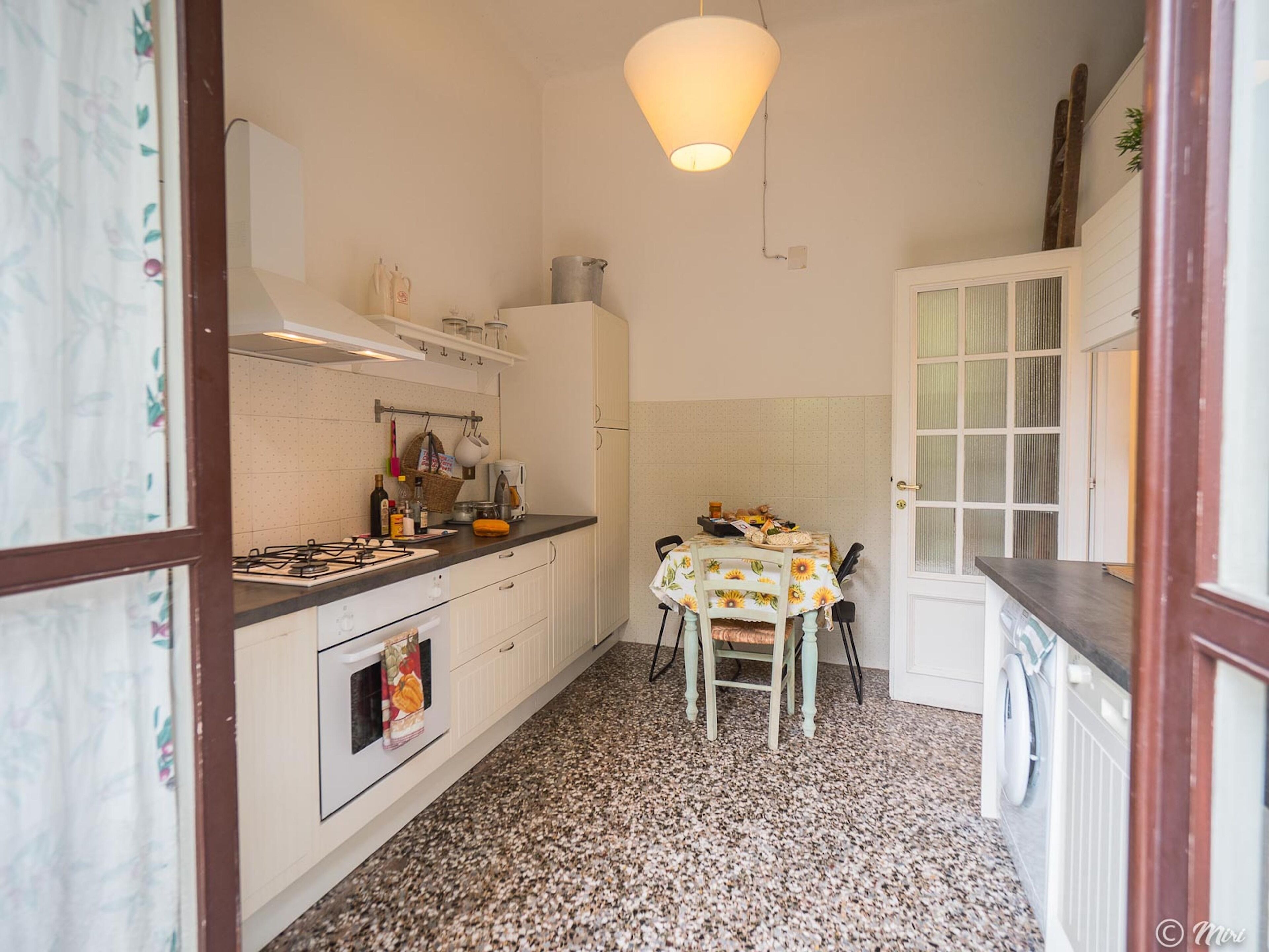 Een keuken met een gasfornuis, oven en een tafel met stoelen. Boven de tafel hangt een lamp.