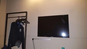 Une pièce avec un téléviseur fixé au mur, un portant à vêtements avec une veste, et une petite table avec une lampe.