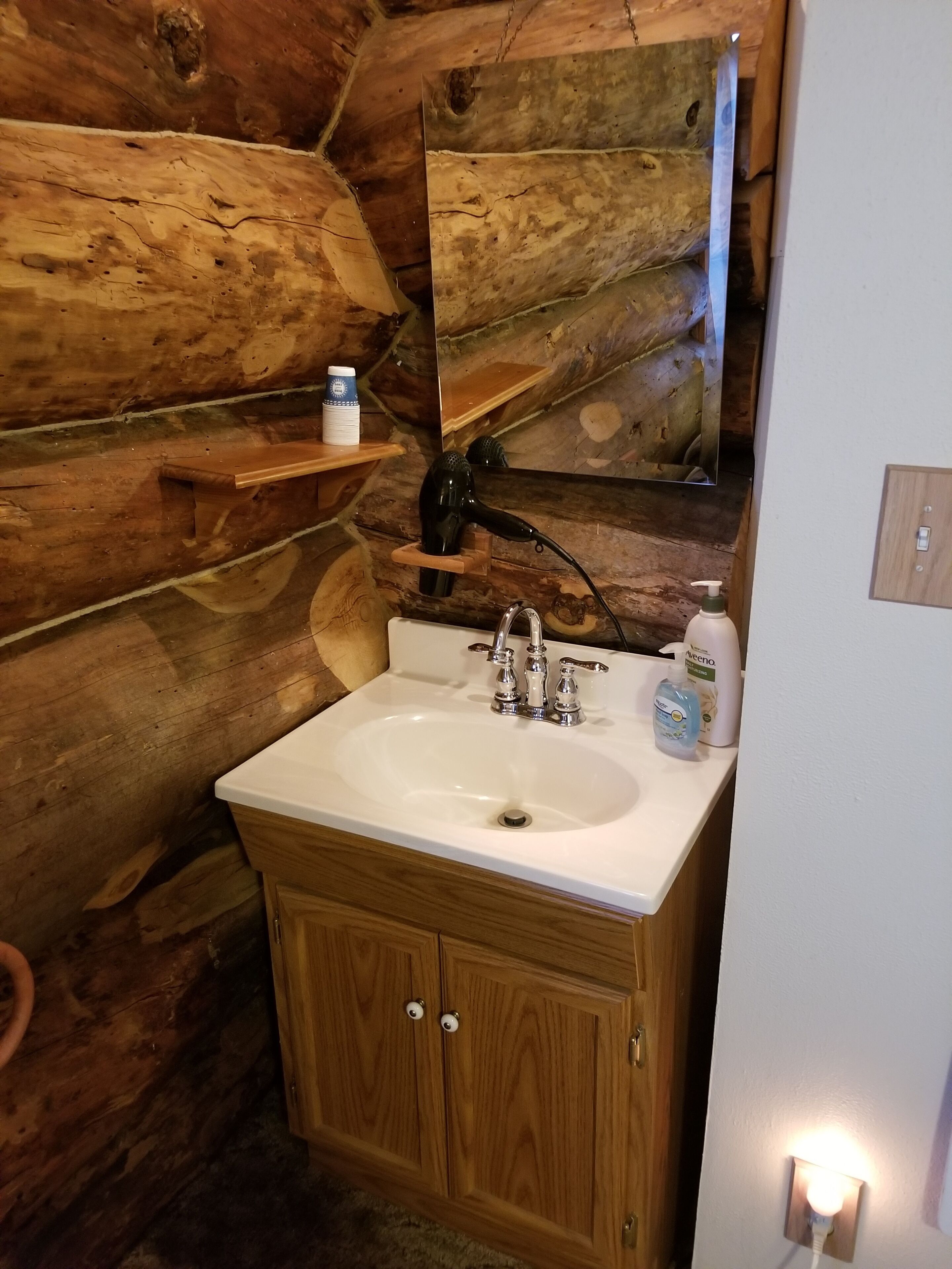 A bathroom with a wooden vanity, a sink, and a mirror. There are toiletries on the vanity and a shelf above the mirror.