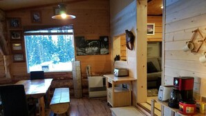 A cozy cabin interior with wooden walls, a dining table, chairs, a coffee maker, and a window overlooking a snowy forest. - Northern Sky Lodge (Fairbanks)