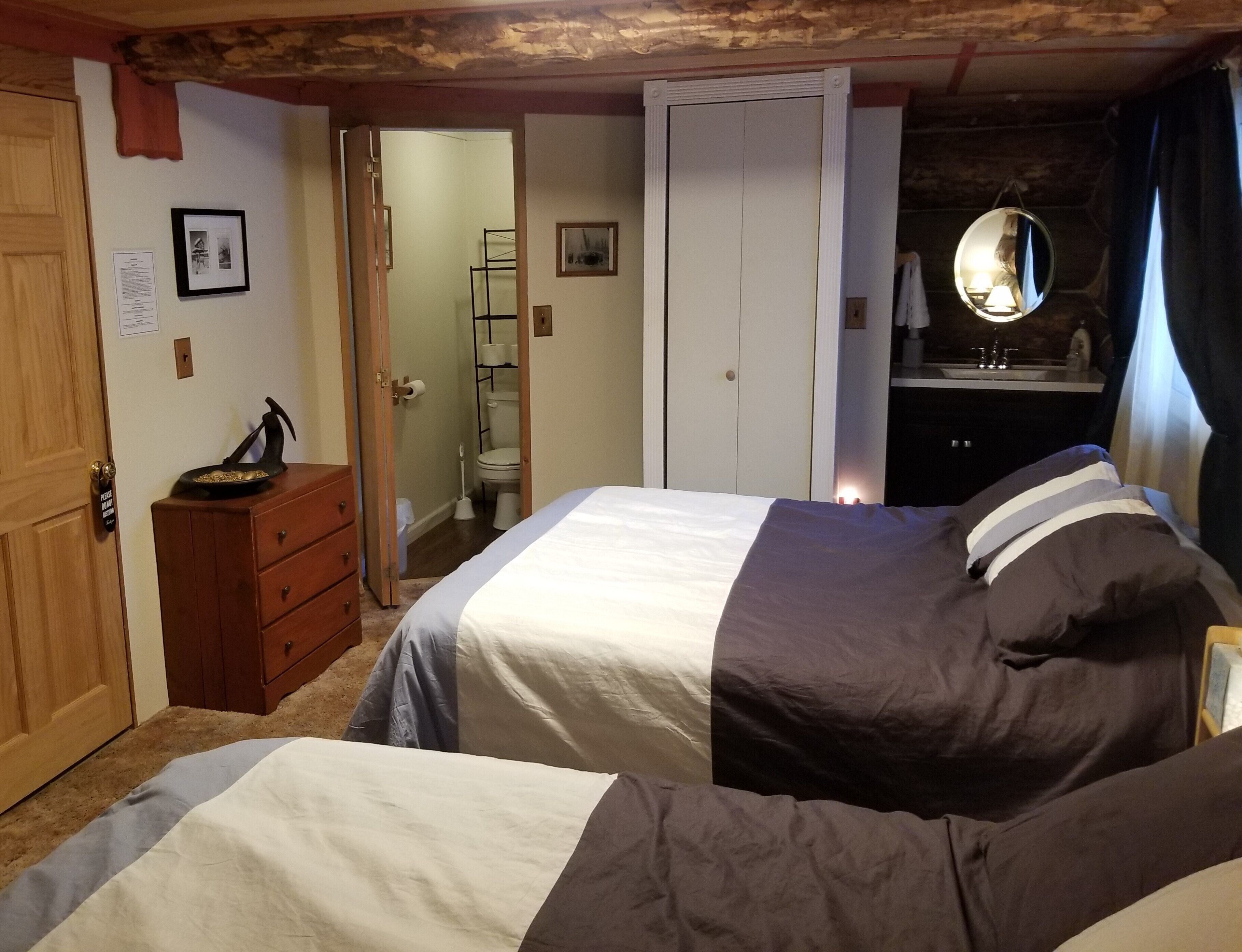 Un dormitorio con dos camas, un aparador de madera, un espejo y un baño visible a través de una puerta abierta.