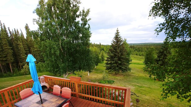 A wooden deck with a table and chairs, surrounded by a wooden railing, overlooking a grassy landscape with trees. - Northern Sky Lodge (Fairbanks)