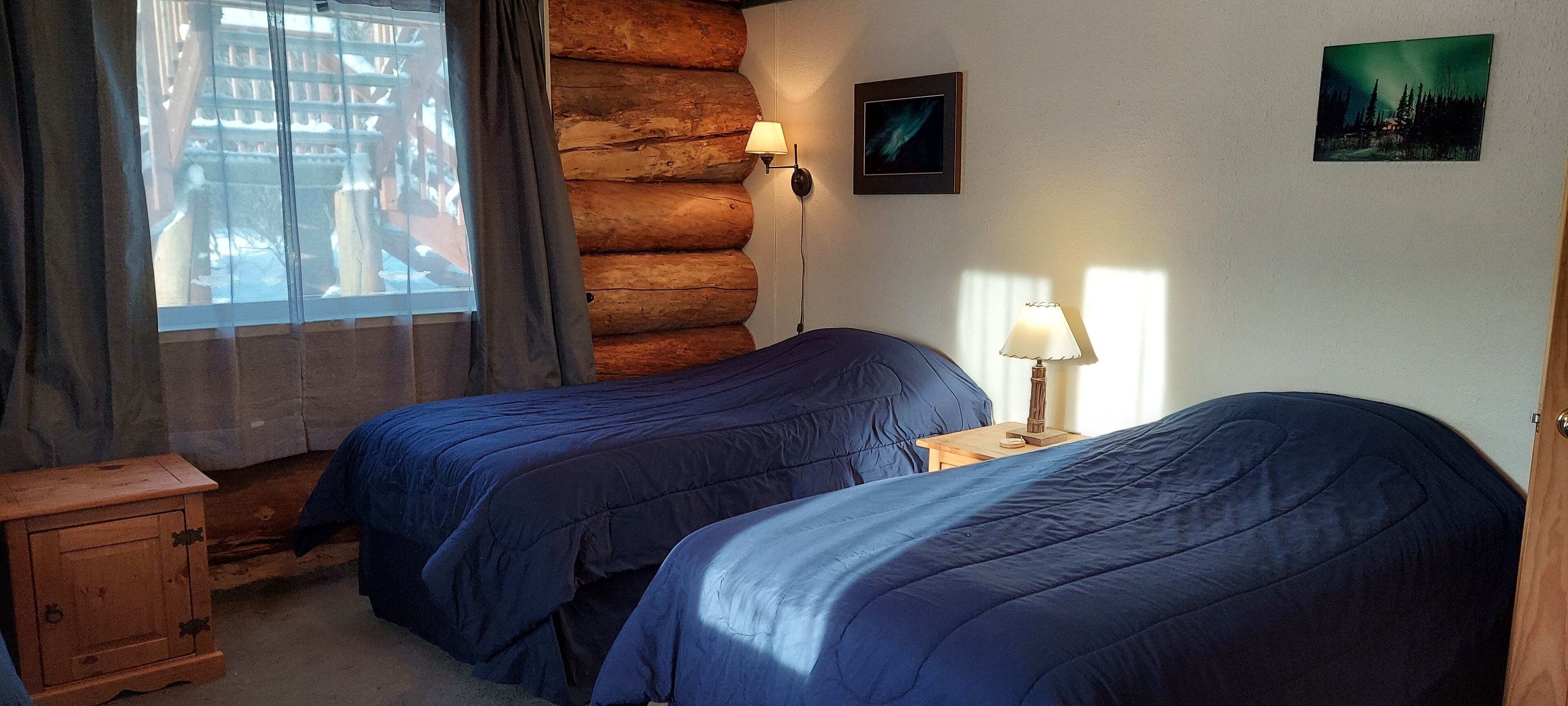A room with two beds, a wooden cabinet, a window with curtains, a wall-mounted lamp, and a framed picture.