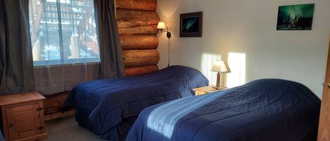A room with two beds, a wooden cabinet, a window with curtains, a wall-mounted lamp, and a framed picture.