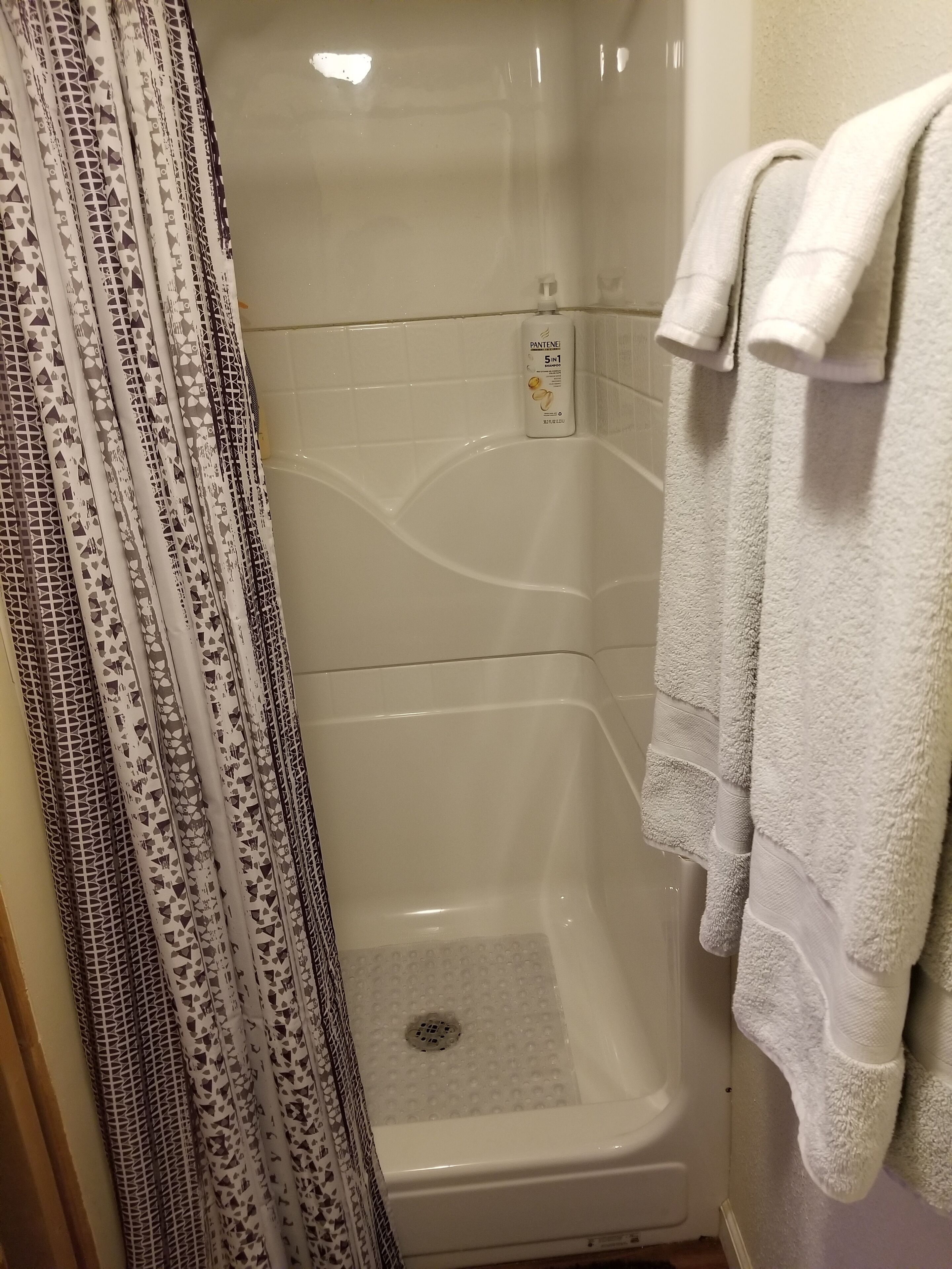 A shower area with a white shower curtain, a white towel rack with towels, and a bottle of Pantene shampoo.