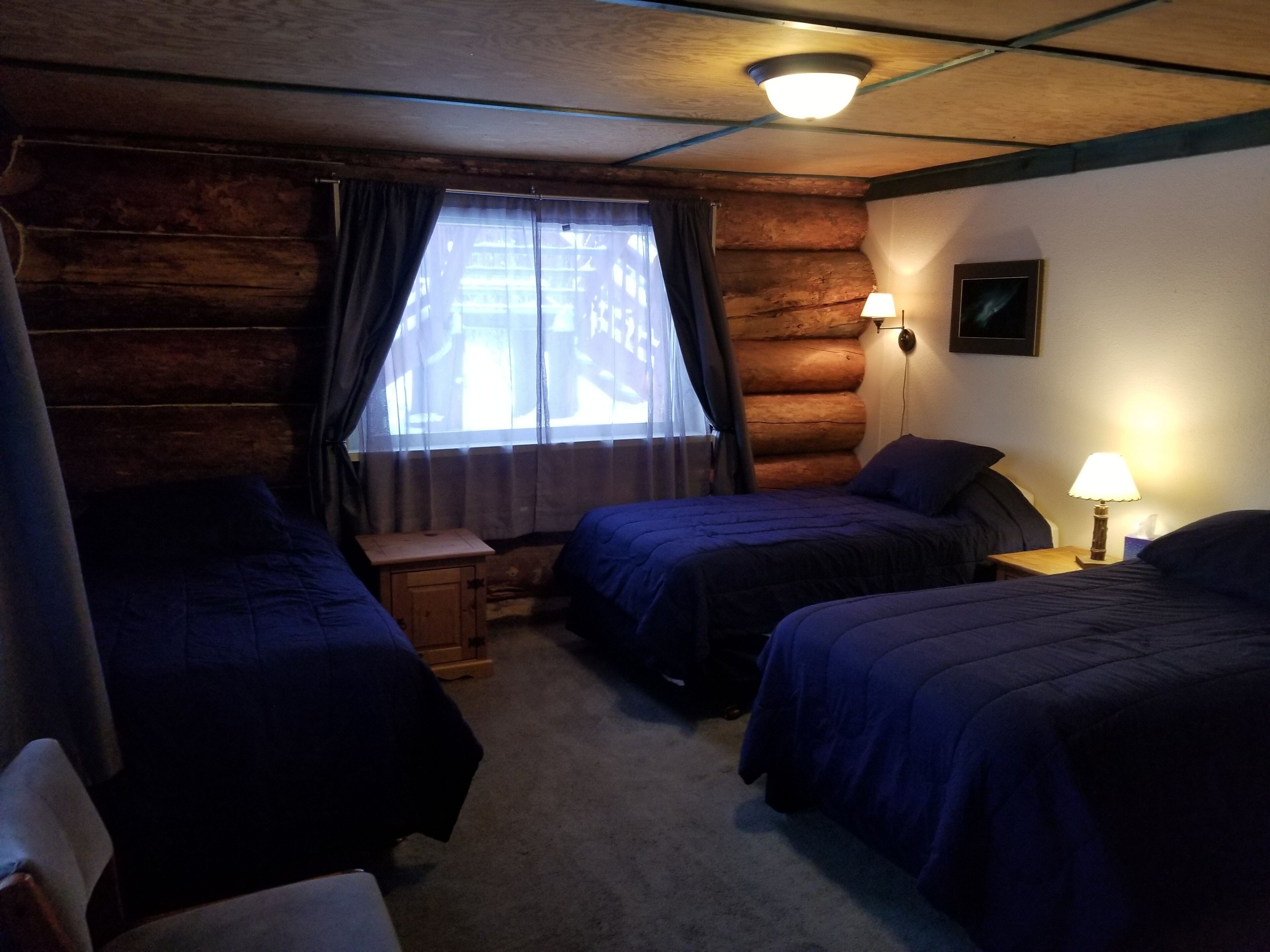 Ein blockhaushaltsähnliches Zimmer mit zwei Betten, einem kleinen Holz-Nachttisch, einer Lampe und einem Fenster mit Vorhängen.