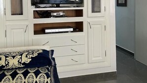 Een woonkamer met een televisie op een witte tv-meubel, een bed met een gekleurd kussen, en een gang die naar een andere kamer leidt.