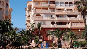 A multi-story hotel with balconies, palm trees, and a mountainous backdrop.