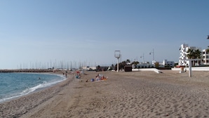 A wide sandy beach with a few people and beach umbrellas. There are buildings and a lifeguard tower in the background.