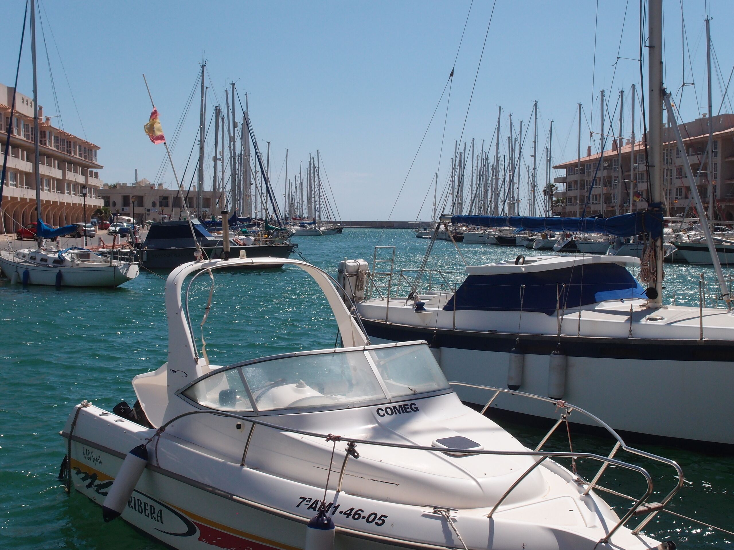 A marina with multiple boats docked, a clear sky, and buildings in the background.