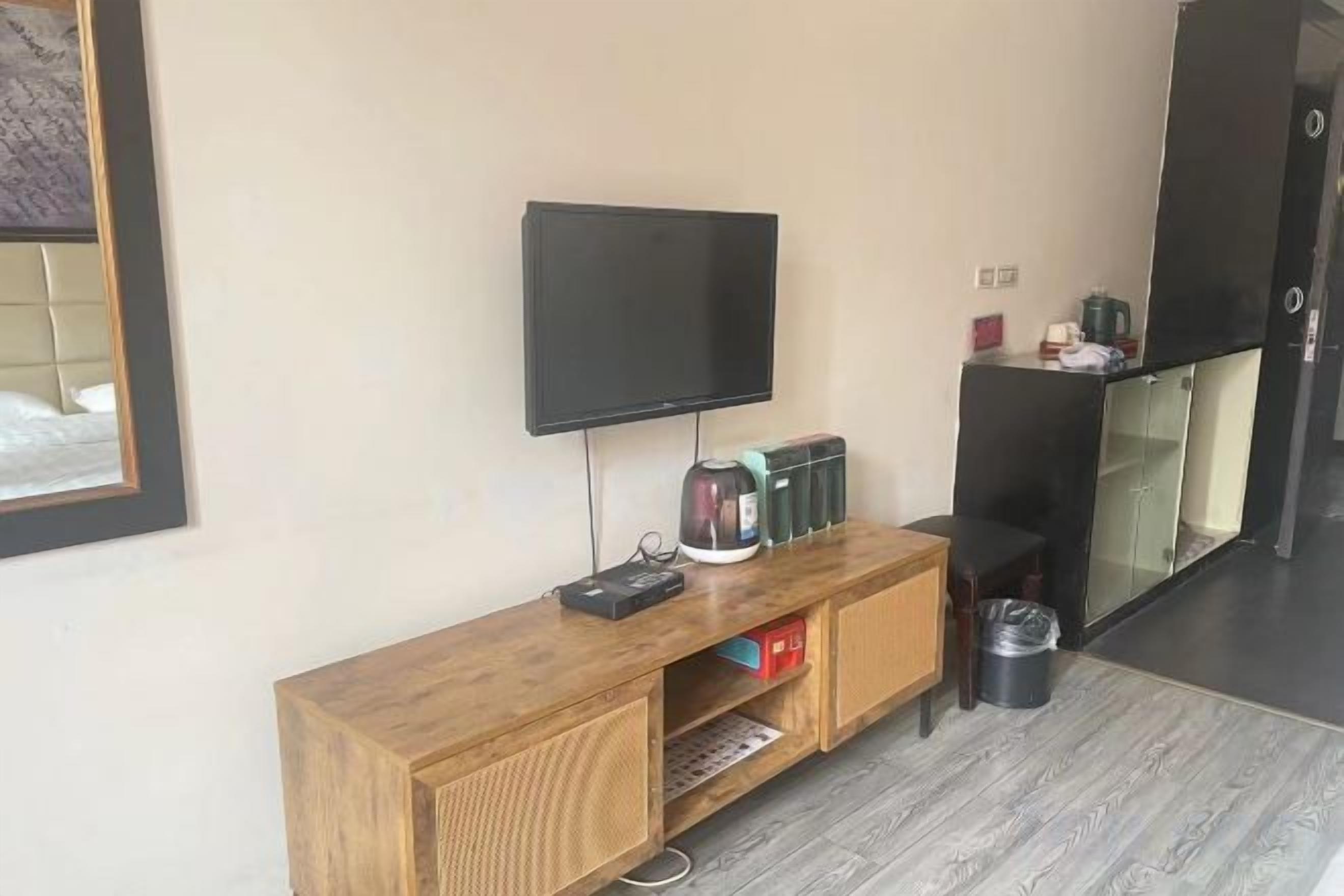 Una habitación con una televisión de pantalla plana montada en la pared, una mesita consola de madera con una tetera, un cubo de basura y un pequeño estante.