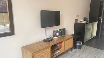 Una habitación con una televisión de pantalla plana montada en la pared, una mesita consola de madera con una tetera, un cubo de basura y un pequeño estante.
