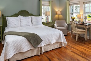 A neatly made bed with a green throw blanket, a chair, a desk with a lamp, and a window with curtains. - The Addison on Amelia Island (Fernandina Beach)