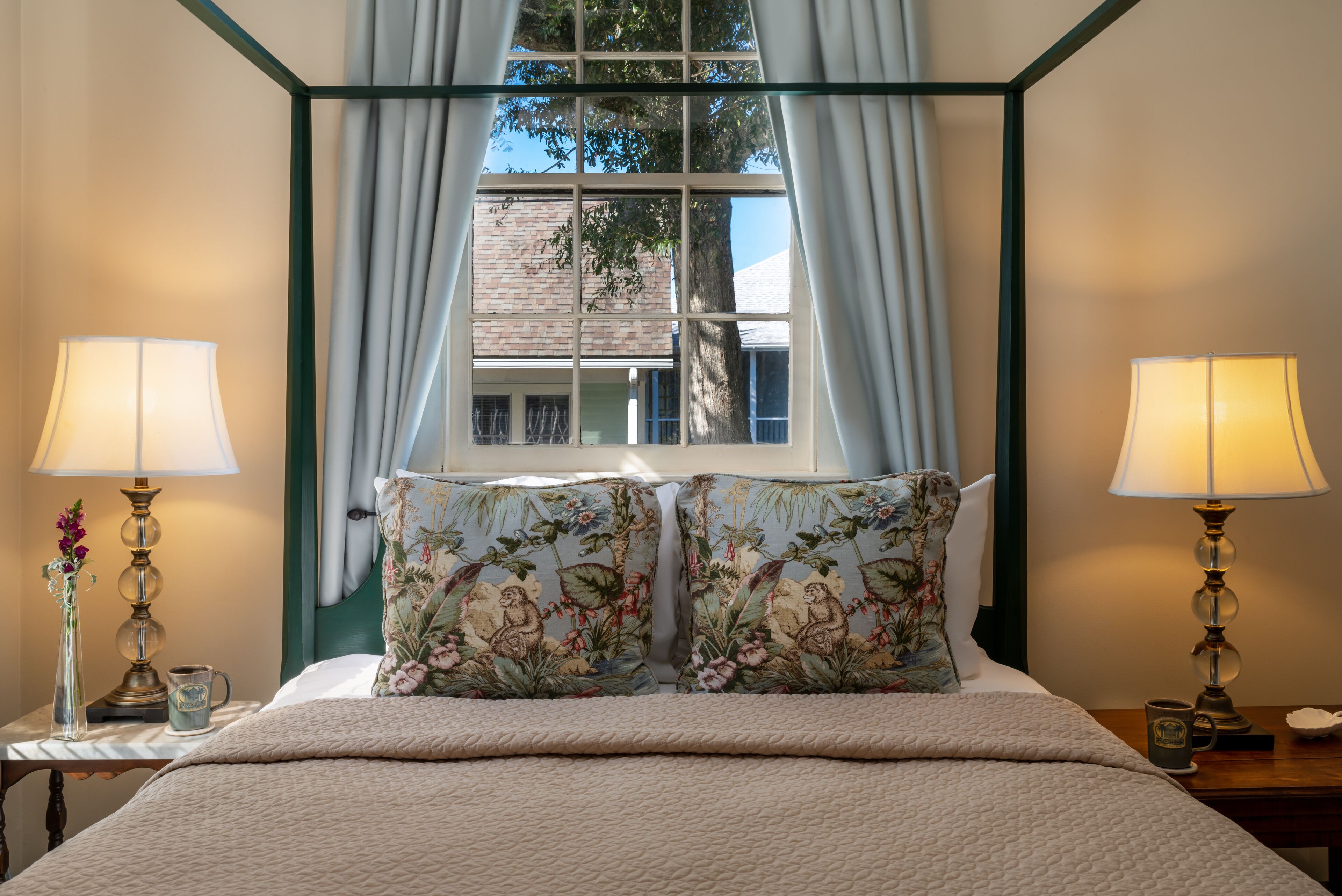 Un lit bien fait, agrémenté de coussins à motifs floraux, deux lampes de chevet et une vue sur un bâtiment, visible à travers une fenêtre ornée de rideaux.
