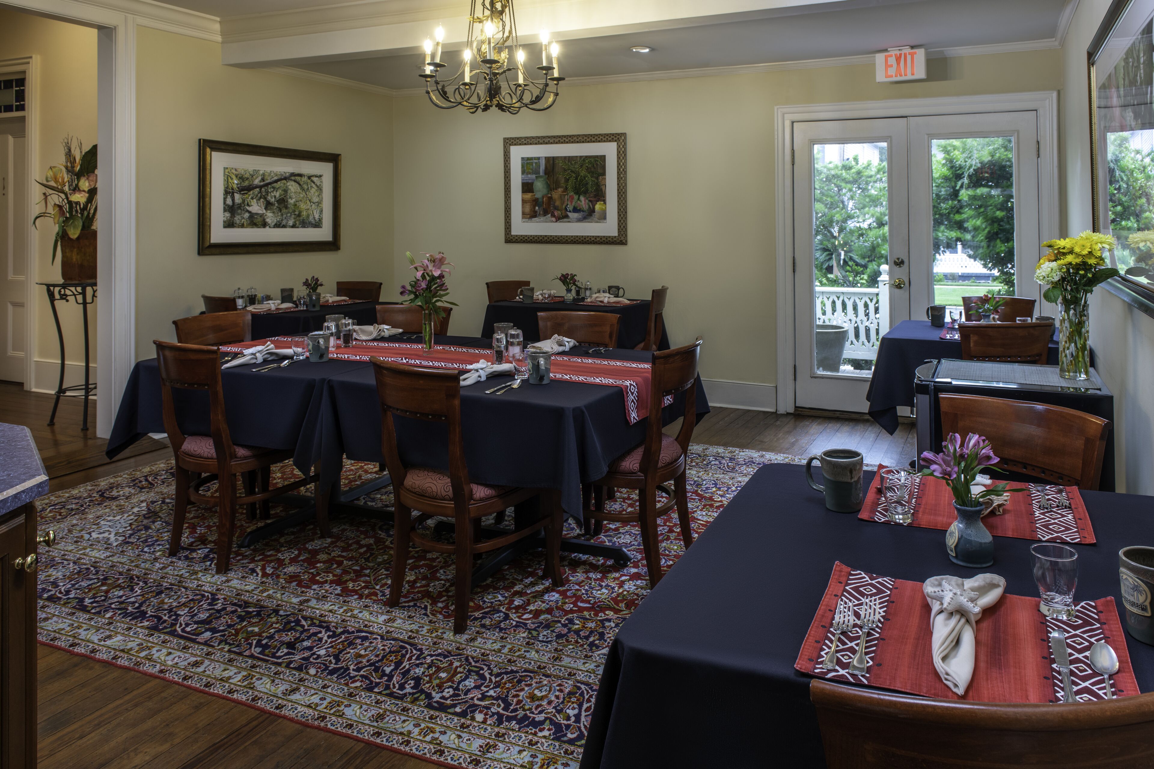 A dining area with tables set for six, featuring dark tablecloths, wooden chairs, and a patterned rug.