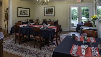 A dining area with tables set for six, featuring dark tablecloths, wooden chairs, and a patterned rug.