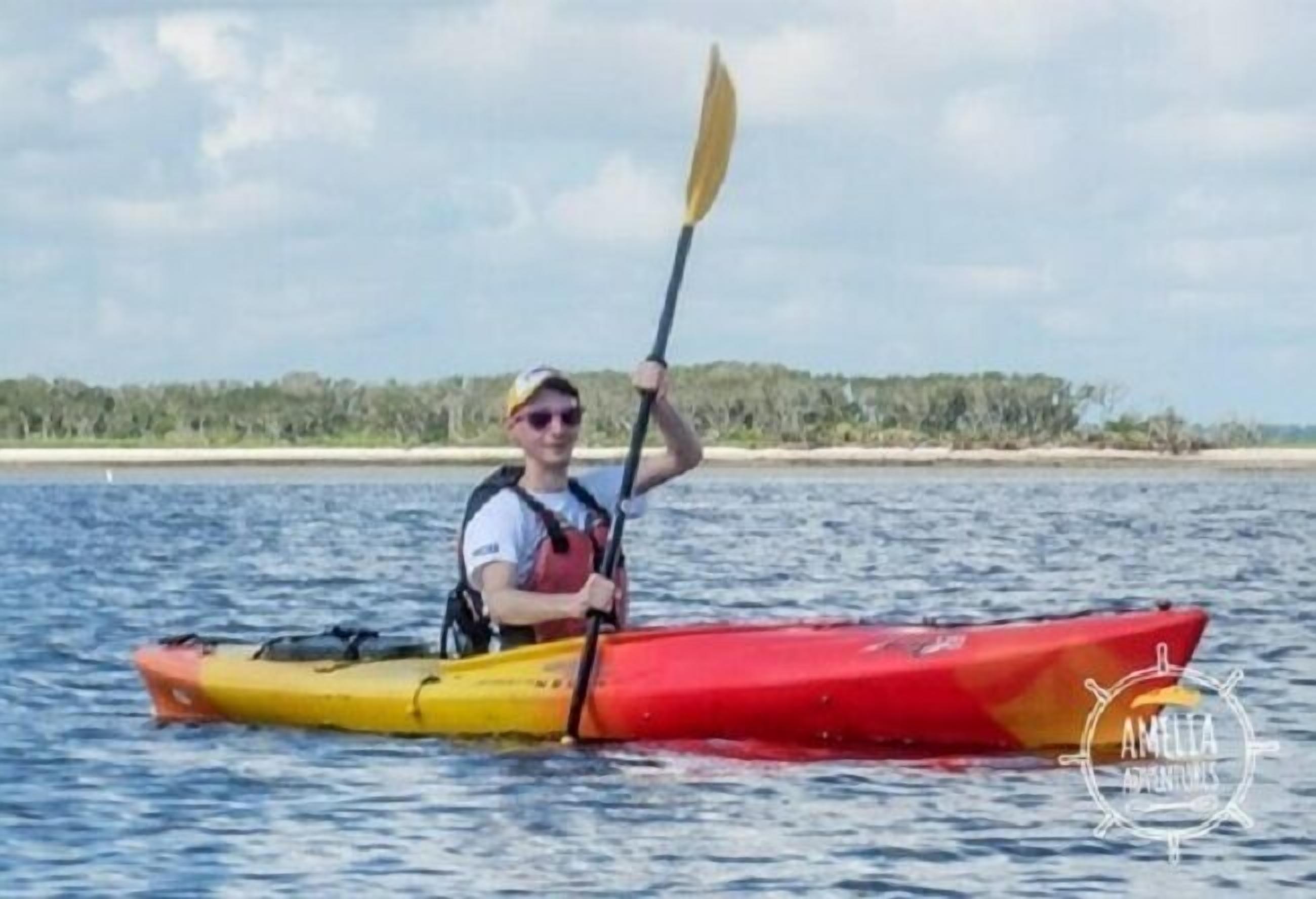 A person in a kayak on a body of water with a beach and vegetation in the background.