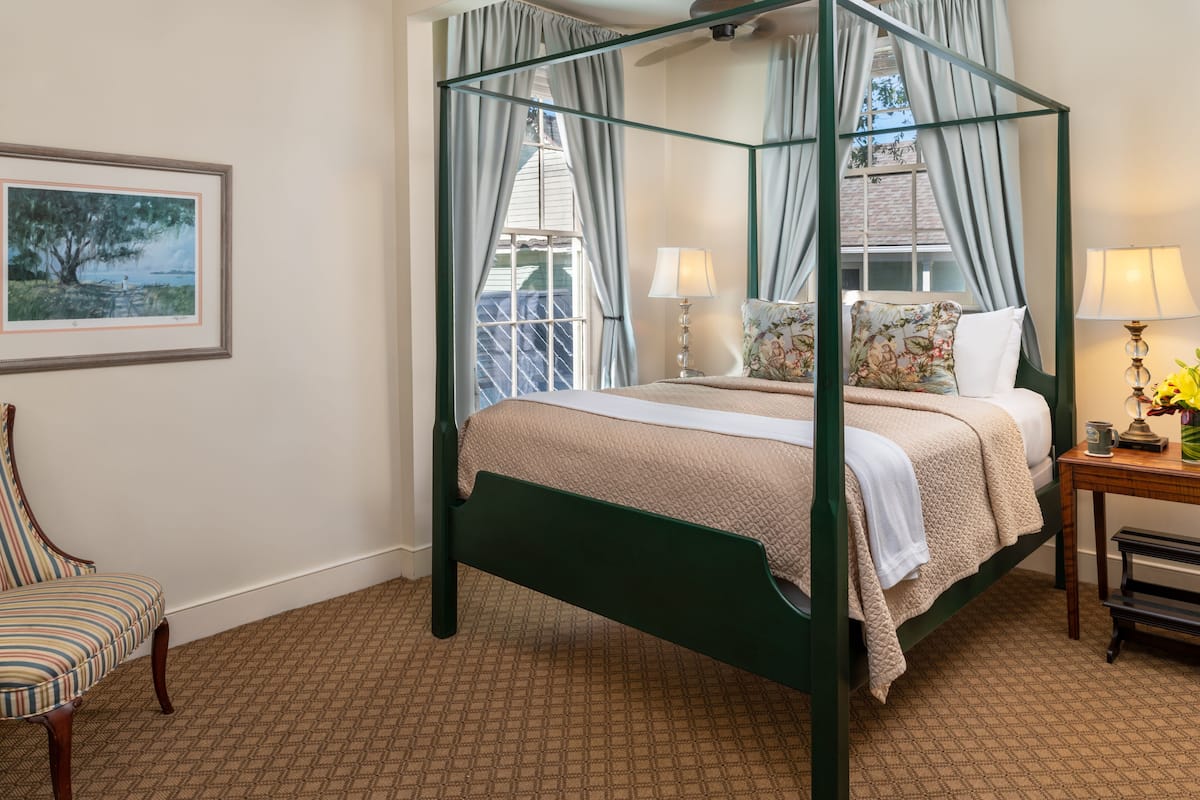 A four-poster bed with a canopy, a chair, a bedside table with a lamp, and a framed picture on the wall.