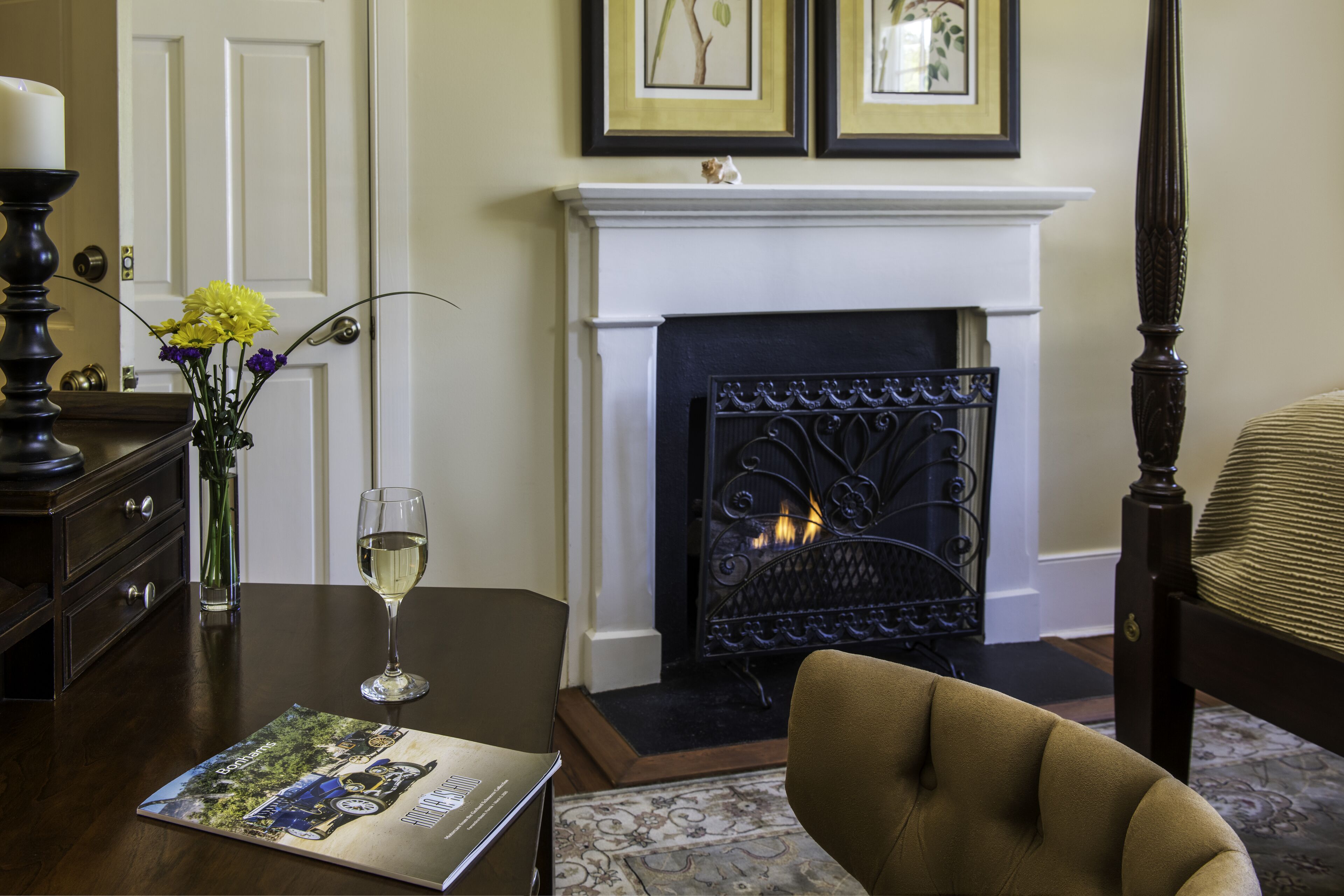 A room with a fireplace, a table with a glass of wine, a vase with flowers, and a framed picture on the wall.