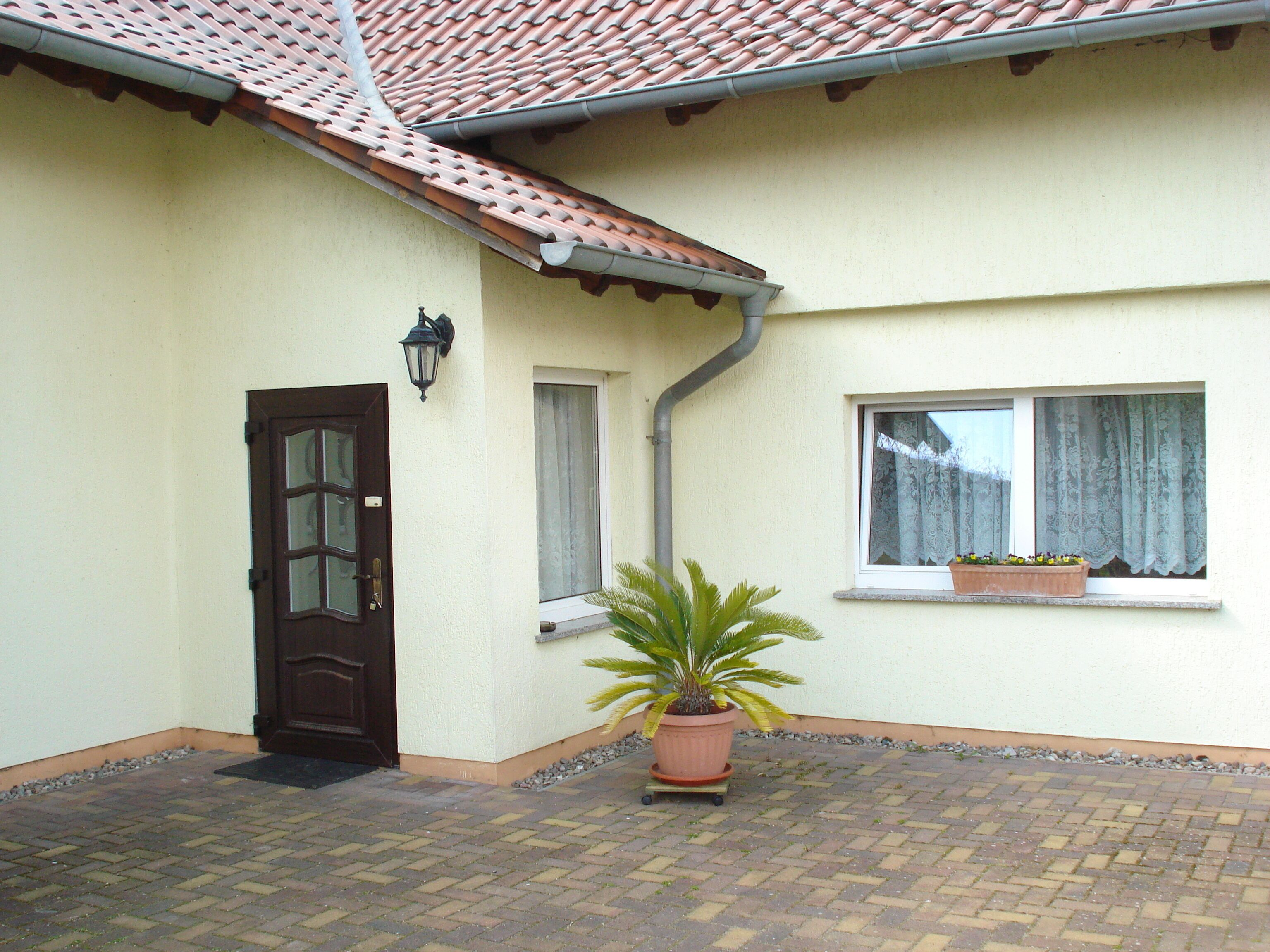 Een huis met pannendak, een deur, een raam met gordijn en een bloempot voor.