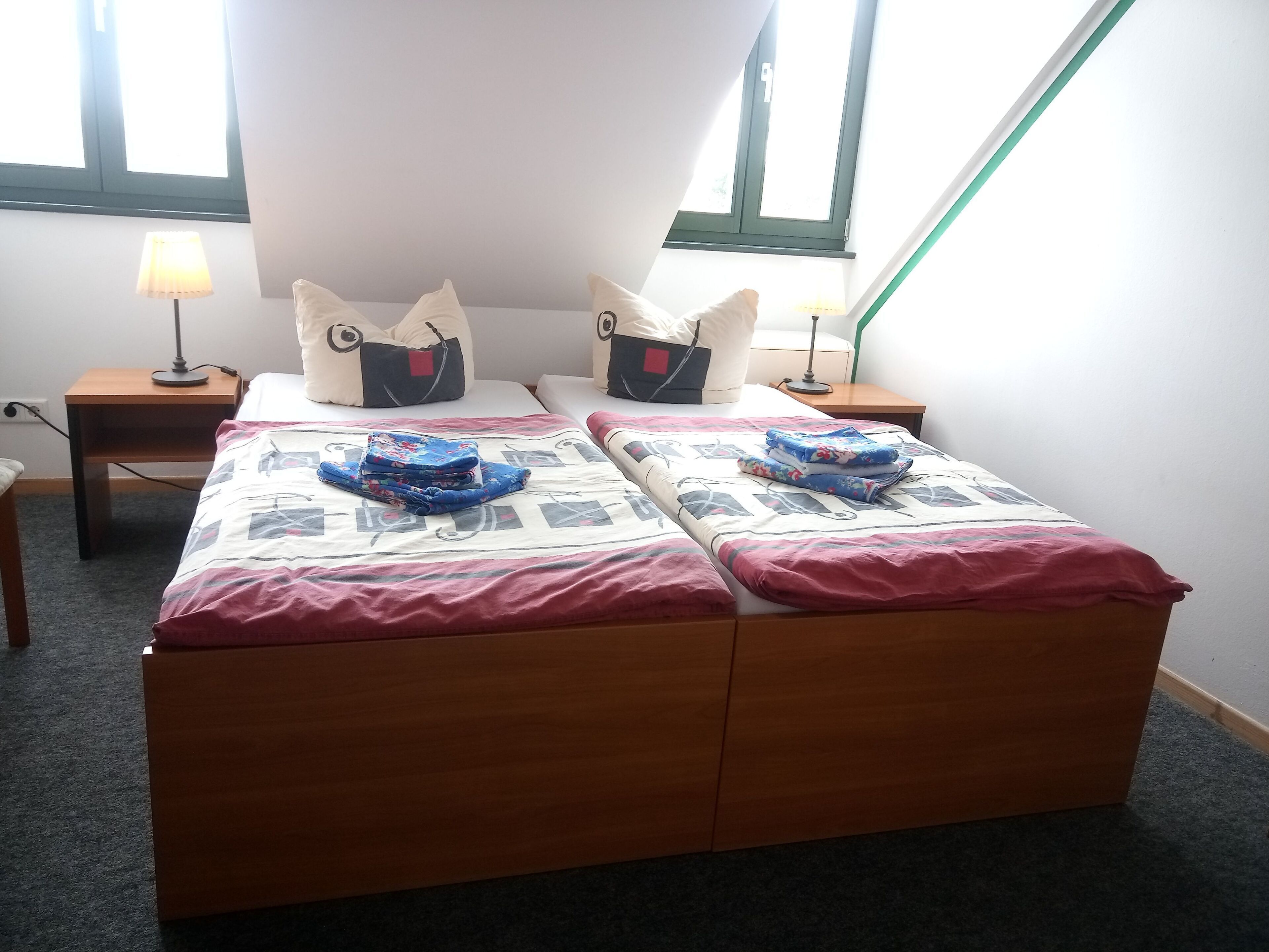 A double bed with a patterned bedspread, two pillows with a unique design, a bedside table with a lamp, and a window with a view.