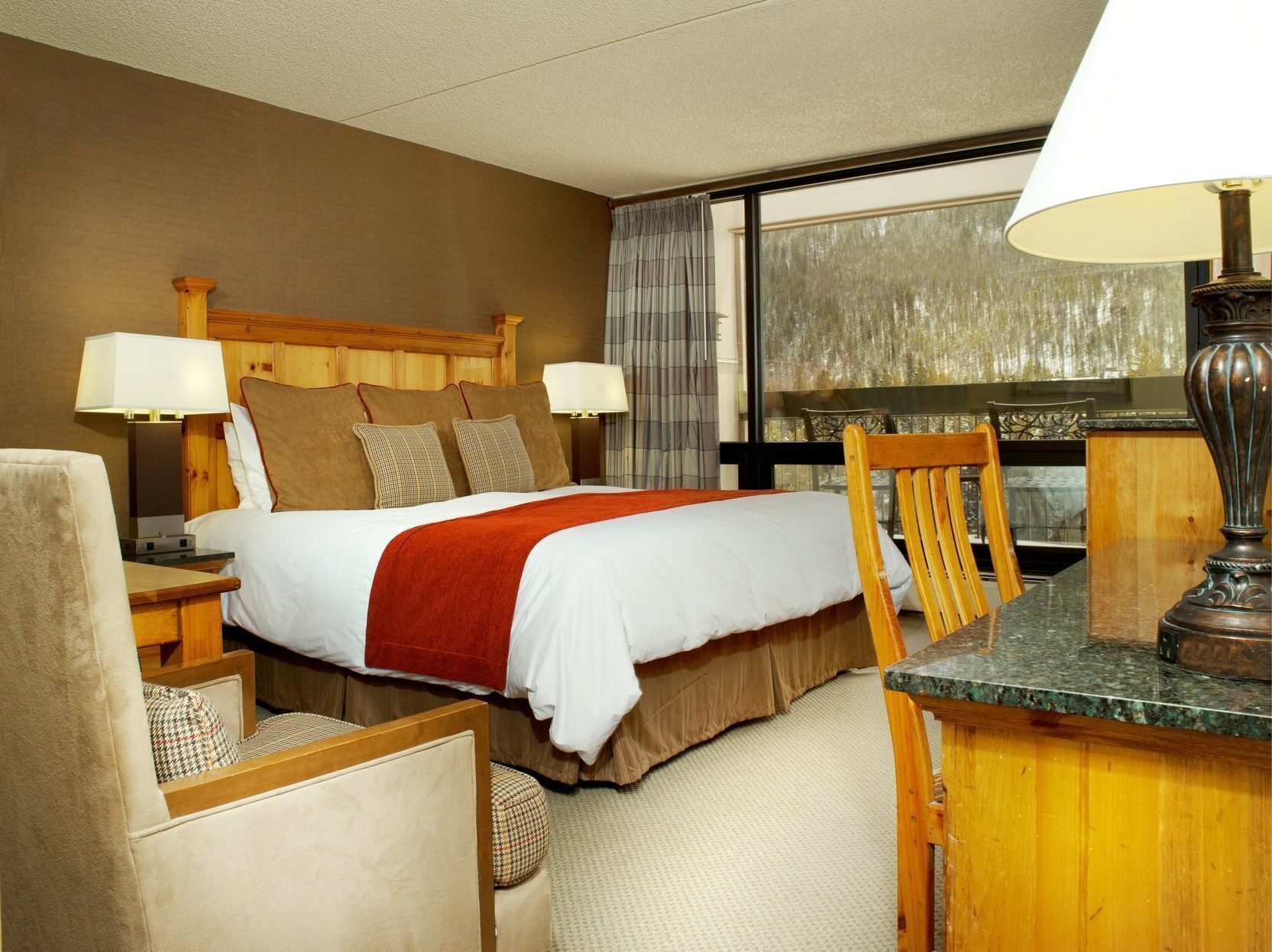 A hotel room with a bed, two bedside lamps, a wooden headboard, a window with curtains, a wooden chair, a table with a lamp, and a carpeted floor.