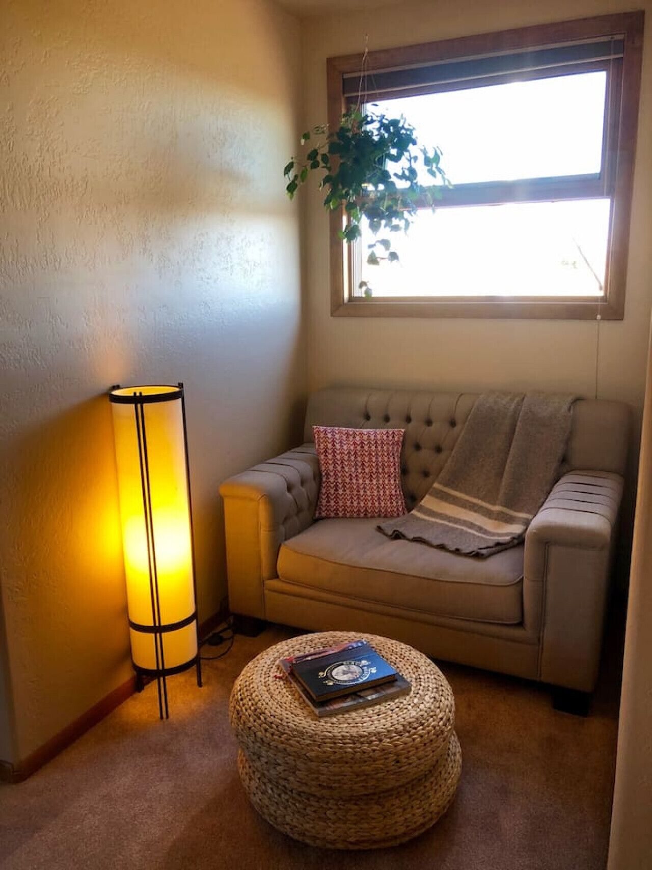 A cozy living room with a sofa, a lamp, a window with a plant, and a round ottoman with a book.