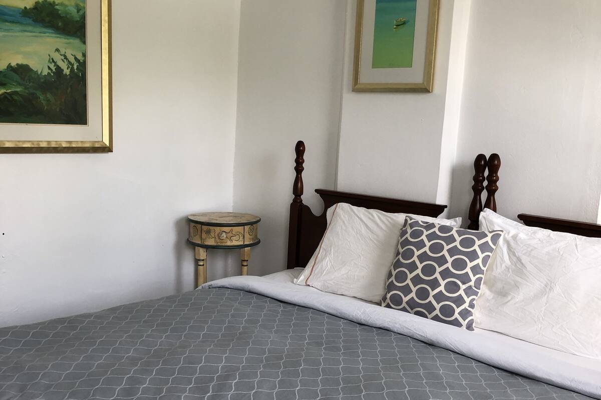 A neatly made bed with a grey patterned bedspread and white pillows, a wooden headboard, and a small round table with a decorative item beside it.