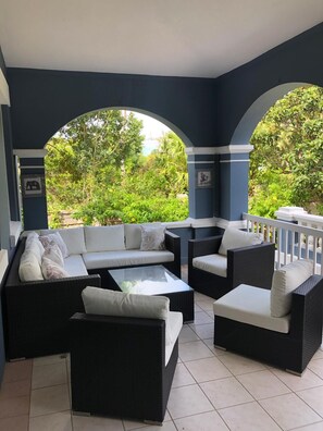 A covered outdoor seating area with wicker furniture and white cushions, overlooking a lush garden. - Bermuda Villa Guest House (Warwick Parish)