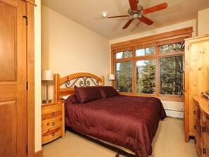 Une chambre à coucher comprenant un lit, une tête de lit en bois, des tables de chevet, une commode, un ventilateur de plafond et une fenêtre donnant sur des arbres.