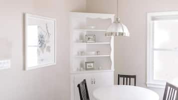 Un coin repas avec une table et des chaises blanches, un luminaire suspendu, un meuble blanc avec des étagères et un tableau encadré au mur.