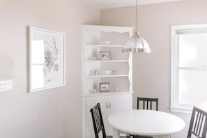Ein Essbereich mit einem weißen Tisch und Stühlen, einer Pendelleuchte, einer weißen Schrank mit Regalen und einem gerahmten Bild an der Wand.