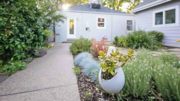 Un sendero conduce a una casa de fachada blanca, rodeada de follaje y un jarrón blanco con plantas.