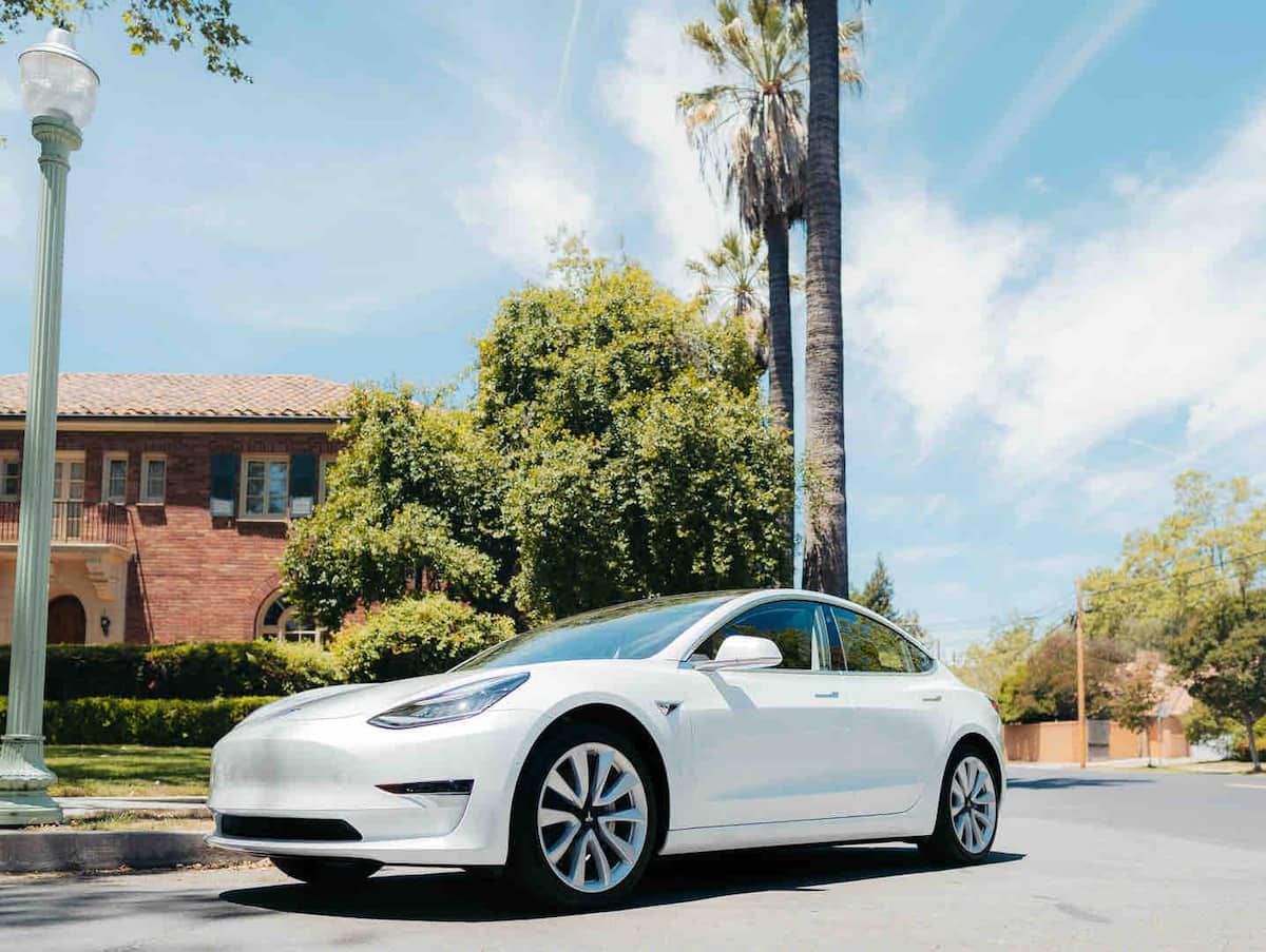 A white Tesla Model S parked on a street with a brick building and palm trees in the background.