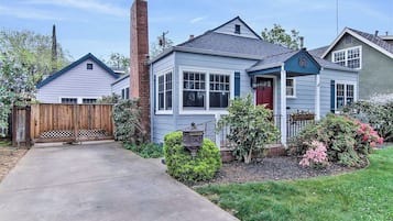 Une maison à deux étages avec un porche à l’avant, une cheminée et un jardin bien entretenu où poussent différentes plantes et fleurs.