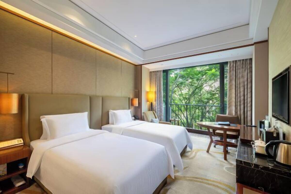 Ein Hotelzimmer mit zwei Betten, einem Schreibtisch, einem Stuhl, einem Fernseher und einem Balkon mit Blick auf Grünflächen.