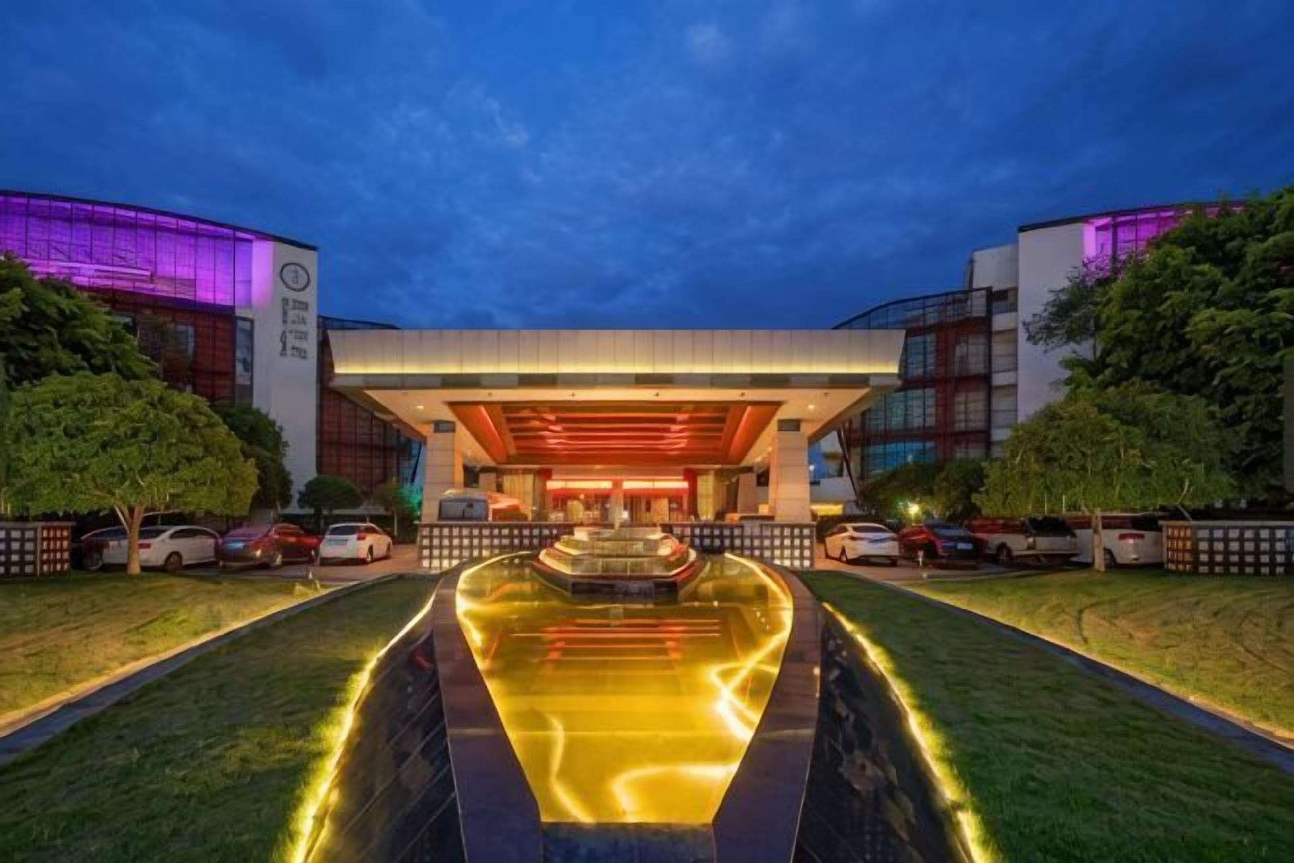 Entrada de hotel con fuente de agua, pasillo cubierto y fachada de edificio moderno.