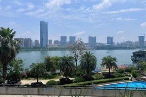 Paisagem urbana com um arranha-céu imponente, um corpo d'água e área de piscina com palmeiras.
