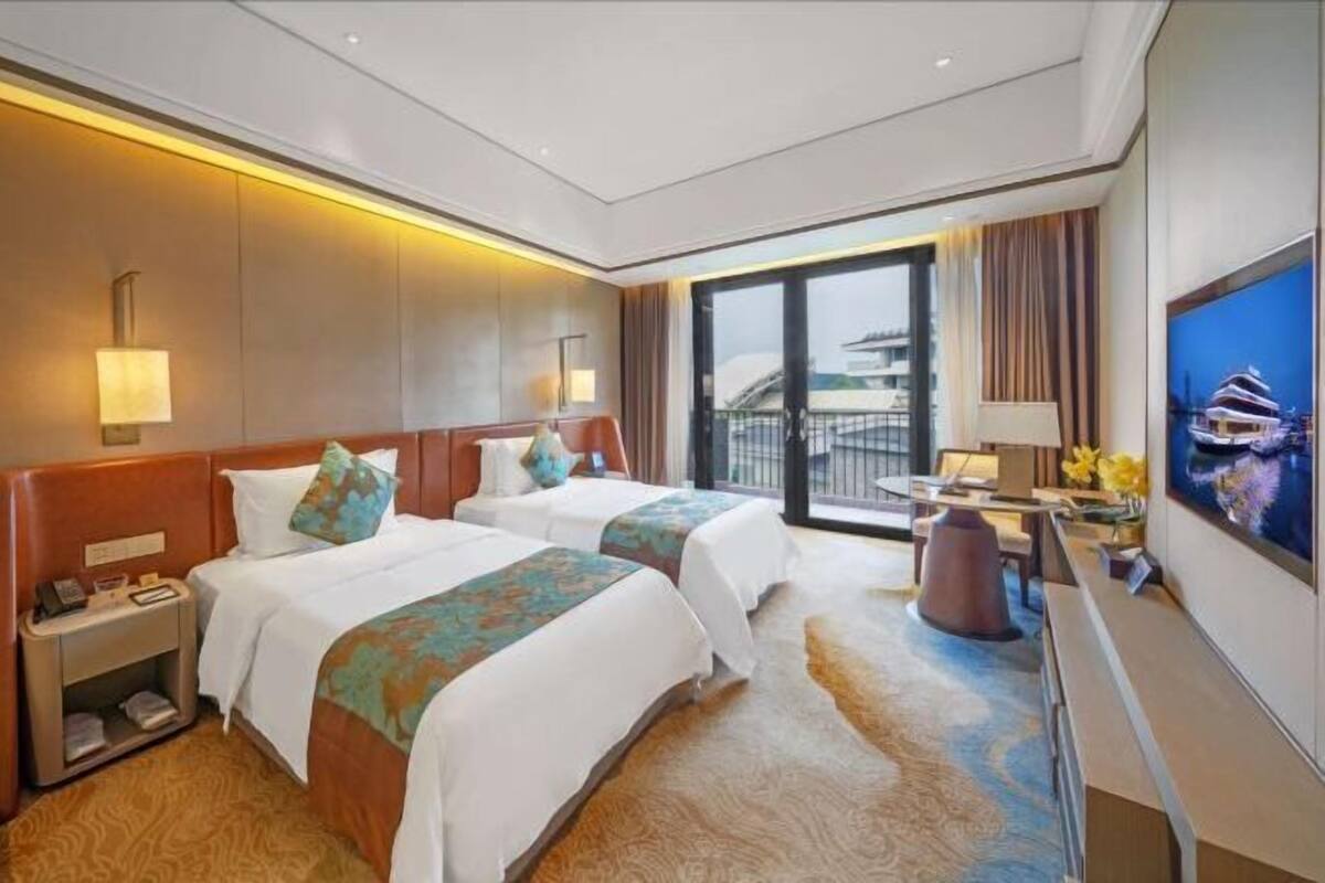 Ein Hotelzimmer mit zwei Betten, einem Schreibtisch, einem Fernseher und Blick auf ein Gebäude.